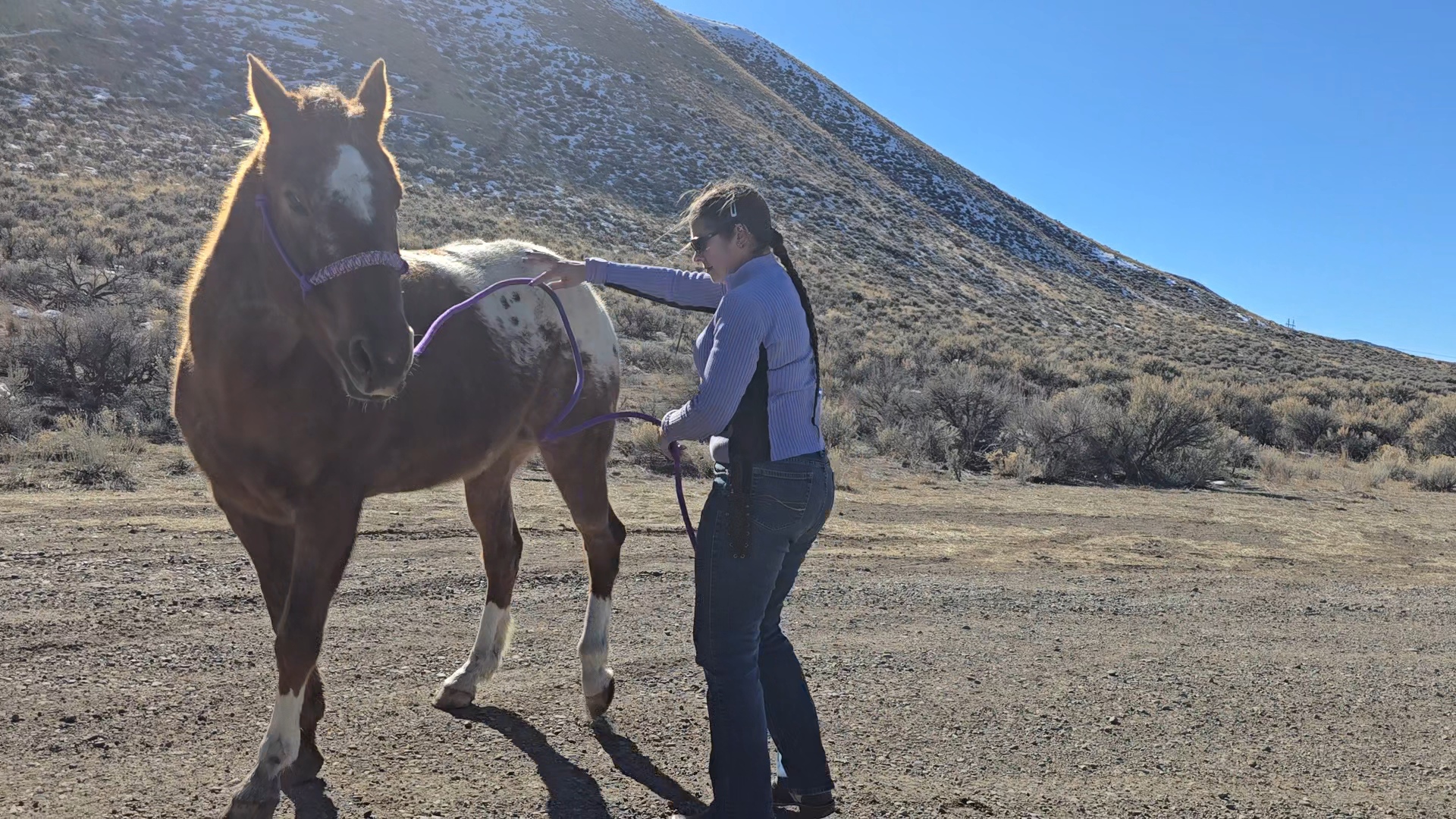 A woman gently interacting with a brown and white horse in a rural outdoor setting, with hills in the background under a clear blue sky.